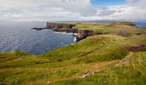A tiny figure in a yellow rain jacket standing in the distance on the cliff edge of a green island. The steep cliffs of the island are visible in the background.